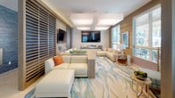 This thoughtfully designed single-floor amenity space blends contemporary coastal aesthetics with functional communal areas. The open-plan layout is anchored by a large communal dining table surrounded by mid-century modern blue upholstered chairs, illuminated by sleek linear pendant lighting above polished light tile flooring. A dedicated lounge zone features generously sized cream sectional sofas accented with warm orange pillows, arranged around dual wall-mounted flat-screen televisions. A fluid, ocean-inspired area rug grounds the space, while wood-slat louvered partitions elegantly divide seating areas, adding texture and visual interest. Bold, multicolored geometric patterned wallcovering in one alcove creates a vibrant nook with built-in bench seating, red accent cushions, and organic-shaped stools. Large windows throughout flood the interior with abundant natural light and frame views of the outdoor pool area and lush landscaping with palm trees. A well-appointed kitchenette offers wood-grain cabinetry, a stone-topped counter with sink, and stainless steel appliances, providing convenient service functionality. The overall ambiance is warm, inviting, and resort-inspired, combining stylish design with a welcoming communal atmosphere.