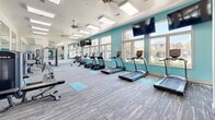 Located at 2002 Stambuk Ln in Statesboro, GA, this fitness center offers a generously sized, single-floor layout designed to accommodate a full range of workout activities. The space is defined by its vibrant teal accent walls, crisp white ceilings, and neutral multi-toned carpet flooring accented with teal exercise mat zones, creating an energetic yet clean aesthetic throughout.

Natural light floods the interior through a row of large windows along one wall, providing views of the surrounding outdoor landscape and lending the space an open, airy feel. Skylight panels and ceiling fans overhead further enhance ventilation and brightness. Mounted flat-screen TVs are positioned throughout for entertainment during workouts.

The cardio area is well-stocked with treadmills, elliptical trainers, and stair climbers arranged to maximize floor space and sightlines to the windows. The strength training section features multi-function cable machines, weight benches, and resistance equipment, with full-length wall mirrors amplifying the sense of space and supporting proper form. A built-in shelving nook along one teal-painted wall stores stability balls and accessories. The overall ambiance is modern, well-lit, and functionally organized.