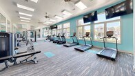 Located at 2002 Stambuk Ln in Statesboro, GA, this fitness center offers a generously sized, single-floor layout designed to accommodate a full range of workout activities. The space is defined by its vibrant teal accent walls, crisp white ceilings, and neutral multi-toned carpet flooring accented with teal exercise mat zones, creating an energetic yet clean aesthetic throughout.

Natural light floods the interior through a row of large windows along one wall, providing views of the surrounding outdoor landscape and lending the space an open, airy feel. Skylight panels and ceiling fans overhead further enhance ventilation and brightness. Mounted flat-screen TVs are positioned throughout for entertainment during workouts.

The cardio area is well-stocked with treadmills, elliptical trainers, and stair climbers arranged to maximize floor space and sightlines to the windows. The strength training section features multi-function cable machines, weight benches, and resistance equipment, with full-length wall mirrors amplifying the sense of space and supporting proper form. A built-in shelving nook along one teal-painted wall stores stability balls and accessories. The overall ambiance is modern, well-lit, and functionally organized.