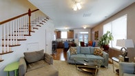 This two-floor home offers a well-connected main level designed for comfortable everyday living. The living room is generously sized, featuring warm earth-toned walls, plush carpeting, and a ceiling fan overhead. Large windows allow natural light to fill the space, which is comfortably furnished with multiple seating areas and a wall-mounted flat-screen television, making it ideal for both relaxing and entertaining.

The open-concept layout connects the living room seamlessly to the dining area and kitchen at the rear of the main floor. The kitchen features warm wood cabinetry, a double-basin sink beneath a window, and ample counter space. A dining area sits adjacent to the kitchen, bathed in natural light from nearby windows and a glass door. A staircase with a classic wood banister and white balusters leads to the upper floor, adding architectural character to the main level. Hardwood flooring in the kitchen and dining area contrasts nicely with the carpeted living room, creating distinct zones within the open floor plan. Throughout the home, indoor greenery and decorative accents contribute to a warm, lived-in ambiance.