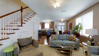 This two-floor home offers a well-connected main level designed for comfortable everyday living. The living room is generously sized, featuring warm earth-toned walls, plush carpeting, and a ceiling fan overhead. Large windows allow natural light to fill the space, which is comfortably furnished with multiple seating areas and a wall-mounted flat-screen television, making it ideal for both relaxing and entertaining.

The open-concept layout connects the living room seamlessly to the dining area and kitchen at the rear of the main floor. The kitchen features warm wood cabinetry, a double-basin sink beneath a window, and ample counter space. A dining area sits adjacent to the kitchen, bathed in natural light from nearby windows and a glass door. A staircase with a classic wood banister and white balusters leads to the upper floor, adding architectural character to the main level. Hardwood flooring in the kitchen and dining area contrasts nicely with the carpeted living room, creating distinct zones within the open floor plan. Throughout the home, indoor greenery and decorative accents contribute to a warm, lived-in ambiance.