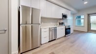 The Nook is a thoughtfully designed single-floor unit that maximizes every square foot with a clean, modern aesthetic. Light wood-tone flooring flows seamlessly throughout the entire space, creating a cohesive and warm atmosphere complemented by crisp white walls and recessed lighting.

The kitchen is a standout feature, outfitted with a full suite of stainless steel appliances including a refrigerator, electric range, dishwasher, and over-the-range microwave. Sleek flat-front white cabinetry extends from floor to ceiling, providing generous storage, while white quartz countertops and a subtle tile backsplash add a polished, contemporary finish.

The living area is bright and open, benefiting from natural light streaming through multiple windows fitted with blinds. A glass-panel entry door connects the space to an outdoor area, enhancing the sense of openness. A sliding-door closet adds practical storage to the living space.

The bathroom continues the modern theme with a white vanity, wood-look tile flooring, and in-unit laundry conveniently tucked behind a folding door. The overall design of The Nook prioritizes functionality and clean lines, making efficient use of its compact footprint while maintaining a bright, welcoming ambiance throughout.