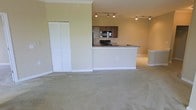 This single-floor space is designed with an open-concept layout that promotes a sense of spaciousness and easy flow between living areas. The expansive carpeted living area provides a comfortable and versatile space, while crown molding along the ceiling adds a refined architectural touch throughout.

The kitchen is positioned at the heart of the space and is well-appointed with dark cabinetry, black appliances including a gas range and built-in microwave, and a peninsula-style breakfast bar that doubles as a casual dining or workspace. Track lighting overhead ensures the kitchen is well-lit and functional.

A set of bifold doors provides access to a generous closet, offering practical built-in storage directly from the main living area. The neutral warm beige wall tones and light carpet create a cohesive and inviting ambiance throughout the space. White baseboards and trim complement the overall aesthetic, adding clean, classic detailing.

Overall, this space balances practicality and comfort, with thoughtful architectural details, efficient use of space, and a layout that suits a variety of living arrangements.