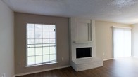 This two-floor space offers a straightforward and functional layout with clean, neutral finishes throughout. The living area serves as a focal point, anchored by a white-painted brick fireplace that extends from floor to a raised stepped hearth, with a recessed niche above the mantel ideal for a television or display. The fireplace adds a classic architectural element that contrasts pleasantly with the otherwise modern, understated aesthetic of the room.

A large picture window floods the living area with natural light, offering a view of the surrounding outdoor greenery. The window's generous size enhances the sense of openness within the space. Neutral warm-gray walls pair well with the rich, dark hardwood flooring, creating a balanced contrast that grounds the room. A textured popcorn ceiling completes the interior overhead, a characteristic detail of the construction period.

The neutral color palette throughout the living area provides a versatile backdrop for a variety of interior design styles. The two-floor configuration of the home suggests additional living and sleeping space upstairs, allowing for clear separation between common areas and private quarters. Overall, the space presents a comfortable, livable environment with notable architectural character from the brick fireplace.