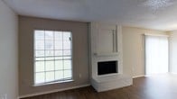 This two-floor space offers a straightforward and functional layout with clean, neutral finishes throughout. The living area serves as a focal point, anchored by a white-painted brick fireplace that extends from floor to a raised stepped hearth, with a recessed niche above the mantel ideal for a television or display. The fireplace adds a classic architectural element that contrasts pleasantly with the otherwise modern, understated aesthetic of the room.

A large picture window floods the living area with natural light, offering a view of the surrounding outdoor greenery. The window's generous size enhances the sense of openness within the space. Neutral warm-gray walls pair well with the rich, dark hardwood flooring, creating a balanced contrast that grounds the room. A textured popcorn ceiling completes the interior overhead, a characteristic detail of the construction period.

The neutral color palette throughout the living area provides a versatile backdrop for a variety of interior design styles. The two-floor configuration of the home suggests additional living and sleeping space upstairs, allowing for clear separation between common areas and private quarters. Overall, the space presents a comfortable, livable environment with notable architectural character from the brick fireplace.