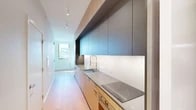 This thoughtfully designed single-floor space showcases a clean, contemporary aesthetic with a cohesive palette of natural materials and neutral tones throughout. The galley-style kitchen is a standout feature, offering floor-to-ceiling cabinetry in a two-tone combination of warm natural wood and matte dark gray, complemented by light stone countertops, an undermount sink with a professional-style pull-down faucet, and a sleek induction cooktop. Under-cabinet lighting enhances the workspace, while a window at the end of the corridor draws in natural light.

The living area is open and airy, distinguished by an exposed concrete structural column that adds an industrial touch against the crisp white walls. Large windows invite abundant natural light, and the space flows seamlessly across wide-plank light hardwood flooring that extends throughout the unit. A set of sliding glass doors connects one of the rooms to an adjoining light-filled space, adding flexibility to the layout.

Additional rooms offer versatility, each featuring the same warm hardwood floors, white walls, and windows with exterior views. The bathroom is finished with large-format light tile, a wall-mounted wood-toned vanity with an integrated white basin, and a glass-enclosed walk-in shower with tile surround, creating a serene and functional space.