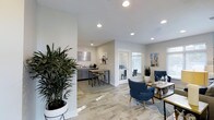 The Aspenwood Apartments leasing office is a thoughtfully designed, single-story space that blends functionality with contemporary style. The open floor plan allows for a seamless flow between distinct areas, creating a welcoming environment for prospective residents and staff alike.

The interior features wood-look tile flooring throughout, lending warmth and a sense of continuity across the space. Recessed ceiling lighting provides consistent, even illumination, complementing the natural light that filters in through white-framed windows. The neutral white and light gray wall palette serves as a clean backdrop, enhanced by tasteful wall art referencing the local community.

A comfortable seating area anchors one side of the space, furnished with deep navy blue accent chairs, a wooden coffee table, and a patterned area rug — ideal for consultations or informal meetings. Adjacent to this, a kitchenette and high-top bar seating area offer a casual space, complete with gray cabinetry and a countertop workspace. A glass-walled private office is visible toward the rear, providing a dedicated area for more formal discussions.

Lush indoor plants are strategically placed throughout, adding a natural, inviting element to the overall modern aesthetic.