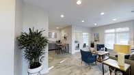 The Aspenwood Apartments leasing office is a thoughtfully designed, single-story space that blends functionality with contemporary style. The open floor plan allows for a seamless flow between distinct areas, creating a welcoming environment for prospective residents and staff alike.

The interior features wood-look tile flooring throughout, lending warmth and a sense of continuity across the space. Recessed ceiling lighting provides consistent, even illumination, complementing the natural light that filters in through white-framed windows. The neutral white and light gray wall palette serves as a clean backdrop, enhanced by tasteful wall art referencing the local community.

A comfortable seating area anchors one side of the space, furnished with deep navy blue accent chairs, a wooden coffee table, and a patterned area rug — ideal for consultations or informal meetings. Adjacent to this, a kitchenette and high-top bar seating area offer a casual space, complete with gray cabinetry and a countertop workspace. A glass-walled private office is visible toward the rear, providing a dedicated area for more formal discussions.

Lush indoor plants are strategically placed throughout, adding a natural, inviting element to the overall modern aesthetic.
