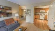 This single-floor residence offers a thoughtfully designed open-concept layout that seamlessly connects the primary living areas. The living room is carpeted and comfortably sized, providing a welcoming space anchored by a stylish sofa and coffee table, with neutral beige walls that create a calm, inviting ambiance throughout.

The dining area flows naturally from the living room, featuring a round dining table with modern metal chairs positioned beneath a pendant light fixture, adding a refined touch to the space. A piece of contemporary wall art bridges the transition between rooms, lending a creative focal point.

The kitchen is accessible via an open archway and features warm-toned wooden cabinetry, dark countertops, and hardwood flooring that contrasts with the carpeted living areas. White appliances, including a range and built-in microwave, are neatly integrated into the cabinetry. Recessed lighting illuminates the kitchen workspace effectively. The overall design balances functionality with comfort, offering a cohesive and practical layout ideal for everyday living. The neutral color palette and consistent design language across all areas contribute to a unified and harmonious aesthetic throughout the home.