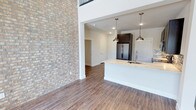 This two-story Frisco residence blends contemporary design with functional living across two well-appointed levels. The main floor makes an immediate impression with a dramatic exposed brick accent wall soaring toward the upper level, complemented by warm wood-look flooring that flows consistently throughout the home. The kitchen is a standout feature, equipped with dark espresso cabinetry extending to the ceiling, stainless steel appliances including a French door refrigerator, granite-style countertops, a mosaic tile backsplash, a built-in wine cooler, and a kitchen island with an undermount sink illuminated by sleek pendant lights. The main living area benefits from tall ceilings and abundant natural light from large windows. A clean entryway with recessed lighting provides a welcoming arrival point. Upstairs, an open hallway with black metal railing overlooks the lower level, enhancing the sense of vertical space. The upper floor includes well-sized bedrooms with ceiling fans, natural light, and closet access. Bathrooms feature dark vanity cabinetry, granite-style countertops, large mirrors, and tiled tub-shower combinations, maintaining a cohesive and polished aesthetic throughout the home.