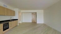This single-floor space showcases a thoughtfully designed open-concept layout that seamlessly integrates the living area with a partial kitchen. The flooring throughout consists of light-toned hardwood planks, lending warmth and a contemporary aesthetic to the interior. White walls and a smooth, neutral ceiling contribute to a bright and airy atmosphere, maximizing the sense of openness across the space.

The kitchen area, positioned to one side, features light wood cabinetry and dark countertops, offering a subtle contrast against the overall pale palette. A structural column near the kitchen defines the boundary between cooking and living zones without fully enclosing either area.

A natural wood-toned entry door serves as a focal point along the far wall, providing a warm architectural detail amid the clean, minimalist surroundings. Recessed ceiling fixtures provide overhead lighting, complementing the overall modern design sensibility. The open floor plan allows for flexible furniture arrangement and versatile use of the living space. The combination of light materials, clean lines, and uncluttered surfaces gives this space a fresh, move-in-ready quality.