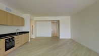 This single-floor space showcases a thoughtfully designed open-concept layout that seamlessly integrates the living area with a partial kitchen. The flooring throughout consists of light-toned hardwood planks, lending warmth and a contemporary aesthetic to the interior. White walls and a smooth, neutral ceiling contribute to a bright and airy atmosphere, maximizing the sense of openness across the space.

The kitchen area, positioned to one side, features light wood cabinetry and dark countertops, offering a subtle contrast against the overall pale palette. A structural column near the kitchen defines the boundary between cooking and living zones without fully enclosing either area.

A natural wood-toned entry door serves as a focal point along the far wall, providing a warm architectural detail amid the clean, minimalist surroundings. Recessed ceiling fixtures provide overhead lighting, complementing the overall modern design sensibility. The open floor plan allows for flexible furniture arrangement and versatile use of the living space. The combination of light materials, clean lines, and uncluttered surfaces gives this space a fresh, move-in-ready quality.