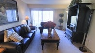 This single-floor space is thoughtfully designed to maximize comfort and functionality. The living room serves as the centerpiece of the home, featuring plush wall-to-wall carpeting that adds warmth underfoot. A large sliding glass door floods the room with natural light and provides direct access to a private balcony, creating a seamless connection between the interior and the outdoors. The living area is generously proportioned, comfortably accommodating a full leather sofa suite and a classic wooden coffee table with elegantly turned legs, which serves as a focal point in the room. A dark entertainment unit anchors one side of the space, offering ample storage and display options. Decorative touches, including potted greenery and vibrant floral arrangements, bring life and color to the interior. Soft, neutral wall tones complement the furnishings and enhance the sense of openness throughout. Wall-mounted lighting fixtures provide a warm ambiance in the evenings, while the natural light from the sliding doors ensures the space feels bright and airy during the day. Overall, this residence offers a cozy yet practical living environment with a classic, timeless aesthetic.