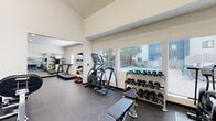 This single-floor fitness center in Hayward, CA (94544) offers a comprehensive workout environment designed to support a wide range of exercise routines. The space is divided into distinct functional zones, each thoughtfully arranged to maximize usability and flow.

The facility features soaring vaulted ceilings in the main strength training area, enhanced by a large ceiling fan that promotes air circulation during workouts. Neutral white walls and rubber-speckled flooring throughout create a clean, professional atmosphere. Large windows fitted with blinds line one side of the cardio zone, allowing natural light to filter in while providing views of the surrounding outdoor area.

Cardio equipment includes treadmills, an elliptical trainer, and a stationary spin bike. A Hoist functional trainer cable machine anchors the strength zone, accompanied by an adjustable weight bench and a full rack of dumbbells spanning a wide weight range. A dedicated stretching and flexibility area houses stability balls, foam rollers, and yoga mats. Full-length mirrors expand the sense of space and assist with form.

A built-in refreshment station features navy blue cabinetry, a marble-style countertop with a sink, floating wood shelves, and a mini-refrigerator. Restroom access is conveniently available on-site.