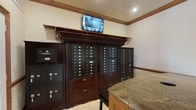This mail and package room is designed with both function and aesthetics in mind. The centerpiece of the space is a substantial built-in dark wood cabinetry unit housing an extensive array of individual mailboxes, providing dedicated mail access for a large number of residents or tenants. Flanking the main unit are secure parcel lockers of varying sizes, ideal for receiving packages of different dimensions safely and conveniently. A ceiling-mounted security monitor positioned above the cabinetry provides continuous surveillance, reinforcing the safety and security of the space. The room features a generously sized counter with a polished marble top, offering a practical surface for sorting and managing mail and deliveries. The interior is finished with warm wood crown molding and baseboards that complement the rich tones of the cabinetry, while recessed lighting and neutral cream-colored walls keep the space bright and welcoming. Light tile flooring completes the clean, professional aesthetic. Overall, the room balances a classic, refined design with highly practical amenities, making it an efficient and attractive hub for mail and package management.