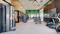 Located at 3461 La Cadena Dr in Riverside, CA, this well-appointed fitness center spans a single floor and offers a comprehensive workout environment across two distinct areas. The main gym floor is generously sized with soaring ceilings, recessed lighting, and large floor-to-ceiling windows that flood the space with natural light and provide views of the surrounding exterior. A bold green accent wall anchors the back of the room, adding a vibrant, energizing aesthetic to the neutral palette. An overhead ceiling fan complements the open, airy feel of the space. The main floor is outfitted with a full row of treadmills and stair climbers, multiple weight benches, a True brand cable and functional trainer system, a dumbbell rack, and additional strength machines. Glass-panel partition walls separate the main gym from an adjacent enclosed studio, which features warm wood-look flooring, floor-to-ceiling mirrors, spin bikes, stability balls, and a dedicated TRX and functional training rig complete with a punching bag, kettlebells, and suspension trainers. Together, these two zones create a versatile, full-featured fitness facility suited for cardio, strength, and group or individual training.