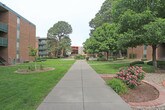 Located at 2201 Ambassador Rd NE in Albuquerque, New Mexico, this single-level outdoor space presents a well-maintained courtyard environment set among classic red brick multi-story buildings. The grounds are defined by a network of wide concrete walkways that provide clear, accessible pathways throughout the property, complemented by dedicated ramp access with metal handrails along the building perimeters.

The landscaping is a notable feature of this space, showcasing a thoughtful mix of lush green lawns, neatly trimmed hedges, flowering rose bushes, decorative gravel beds, and mature trees that provide generous shade across the grounds. Yucca plants and cypress trees add a distinct Southwestern character to the plantings, blending regional flora with traditional garden elements.

Brick-edged garden beds frame curved planting areas, adding structure and visual interest to the open green spaces. The courtyard setting is framed by the surrounding multi-story brick buildings, which feature green-accented balconies overlooking the grounds. Utility areas, including recycling stations and cluster mailboxes, are discreetly positioned along the building edges. The overall environment is open, green, and pedestrian-friendly, creating a welcoming communal outdoor space with a calm and well-kept atmosphere.