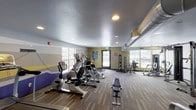 Located at 101 E 88th Ave in Thornton, Colorado, this fitness center is a single-floor facility designed to support a comprehensive workout experience. The space is generously sized, with an open floor plan that accommodates a wide variety of exercise equipment without feeling crowded.

The cardio zone features a robust lineup of True and Matrix brand machines, including treadmills, ellipticals, stationary bikes, and stair climbers, all arranged in a row to maximize floor space. Natural light filters in through large windows along the perimeter walls, offering views of the surrounding area and creating an energizing atmosphere.

The strength training section includes a multi-functional Matrix cable machine, a seated leg press, and various resistance machines suited for both upper and lower body workouts. A dedicated free weight area provides a full dumbbell rack and a flat bench, ideal for a range of lifting exercises.

The flooring consists of durable, striped carpet tiles that define workout zones throughout the room. Recessed lighting and exposed HVAC ductwork contribute to the facility's functional aesthetic. Walls are painted in warm yellow and neutral tones with decorative banding, adding visual interest to the space.

Overall, this fitness center delivers a well-organized, fully equipped environment suitable for a broad range of fitness routines.