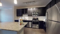 This single-floor residence is thoughtfully designed with a cohesive blend of contemporary finishes and functional layouts. The kitchen serves as a focal point, showcasing dark espresso-toned cabinetry that contrasts elegantly with light granite countertops. A full suite of stainless steel appliances — including a range, built-in microwave, dishwasher, and refrigerator — provides a well-equipped cooking space. A white subway tile backsplash adds a clean, classic detail, while a center island with pendant lighting above offers additional prep space and casual seating potential. Track lighting further illuminates the workspace. Wood-look flooring extends throughout the main living areas, contributing warmth and visual continuity.

The open layout connects the kitchen to the broader living space, with multiple doors visible along one wall suggesting well-organized storage or additional rooms. The bedroom features plush carpeting underfoot and two large windows fitted with blinds, allowing natural light to fill the room while maintaining privacy.

The bathroom is finished with the same granite vanity countertop seen in the kitchen, paired with a dark vanity cabinet, a large mirror, wood-look flooring, and a full soaking tub with tiled surround, creating a clean and cohesive aesthetic throughout.