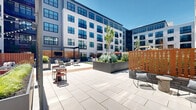 Located at 145 City Ave in Bala Cynwyd, PA, this expansive outdoor courtyard is a beautifully curated amenity space set within a modern multi-story residential building. The courtyard is framed on all sides by the building's contemporary façade, characterized by large windows, private balconies, and a clean mix of white paneling and gray siding that lends the space a polished, urban aesthetic.

At the heart of the courtyard lies a generous stretch of lush green turf, providing an open, inviting area for recreation and leisure. Flanking the turf are wide-format concrete pavers and warm wood decking that define distinct activity zones throughout the space. Multiple wooden pergolas with string lights serve as charming focal points, sheltering intimate seating arrangements that include lounge sofas, accent chairs, and dining sets—perfect for both casual gatherings and quiet relaxation.

A circular wood-tiled deck beneath a curved overhead structure offers a dramatic, design-forward gathering spot. Raised planters filled with ornamental grasses, lush shrubs, and young trees add layers of natural texture and greenery throughout. An outdoor grill station with adjacent bistro seating further enhances the space's functionality. The overall ambiance is modern, welcoming, and thoughtfully landscaped, creating a vibrant communal outdoor retreat.