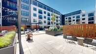 Located at 145 City Ave in Bala Cynwyd, PA, this expansive outdoor courtyard is a beautifully curated amenity space set within a modern multi-story residential building. The courtyard is framed on all sides by the building's contemporary façade, characterized by large windows, private balconies, and a clean mix of white paneling and gray siding that lends the space a polished, urban aesthetic.

At the heart of the courtyard lies a generous stretch of lush green turf, providing an open, inviting area for recreation and leisure. Flanking the turf are wide-format concrete pavers and warm wood decking that define distinct activity zones throughout the space. Multiple wooden pergolas with string lights serve as charming focal points, sheltering intimate seating arrangements that include lounge sofas, accent chairs, and dining sets—perfect for both casual gatherings and quiet relaxation.

A circular wood-tiled deck beneath a curved overhead structure offers a dramatic, design-forward gathering spot. Raised planters filled with ornamental grasses, lush shrubs, and young trees add layers of natural texture and greenery throughout. An outdoor grill station with adjacent bistro seating further enhances the space's functionality. The overall ambiance is modern, welcoming, and thoughtfully landscaped, creating a vibrant communal outdoor retreat.