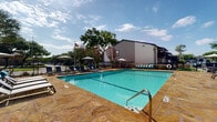 This outdoor amenity space centers around a generously sized rectangular swimming pool with clear turquoise water and defined lane markings. The pool is framed by a broad, natural flagstone deck in warm amber and tan tones, providing ample room for lounging and socializing. Rows of modern reclining chaise lounges with teal accent cushions are arranged along both sides of the pool, offering comfortable spots to relax and soak up the sun. Multiple large patio umbrellas are positioned throughout the deck, providing shaded relief during warm days. A dedicated outdoor dining area features a round table with coordinating chairs and an umbrella, ideal for gatherings. On one end of the pool deck, a covered pergola shelters a built-in grill station and additional seating, creating a functional outdoor kitchen and entertainment zone. The entire pool area is enclosed by a black wrought iron fence, ensuring privacy and security while maintaining open sightlines to the surrounding mature trees and landscaping. An American flag is prominently displayed, adding a patriotic touch to the setting. The combination of durable stone hardscaping, lush greenery, and thoughtfully arranged furnishings creates an inviting and resort-like atmosphere suitable for both relaxation and outdoor entertaining.