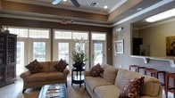 This single-floor space is thoughtfully designed to create a warm and inviting atmosphere. The main lounge area is anchored by a striking tray ceiling accented with crown molding and fitted with a ceiling fan, lending the room a sense of elegance and dimension. Recessed lighting complements the natural light that pours in through a wall of large French doors and transom windows, offering views of an outdoor deck area surrounded by lush greenery.

The interior is finished in rich, earthy tones — warm taupe and brown walls paired with crisp white trim create a refined yet comfortable aesthetic. Two generously sized beige sofas are arranged facing each other around a round pedestal accent table, which is adorned with a lush floral arrangement, making it a natural focal point. Decorative throw pillows in deep jewel tones add layers of texture and color throughout the seating area.

A wall-mounted flat-screen television is positioned for easy viewing, framed by a neutral-toned accent wall. A framed art piece and a bold red side table contribute additional character to the space. The flooring features light-toned tile, partially covered by a woven area rug. Overall, the space exudes a polished, community-oriented ambiance suited for lounging and socializing.