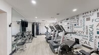 The fitness center at AVIA Lofts at Maple Road is a well-appointed, single-level exercise facility located in Buffalo, NY. Designed with a clean, modern aesthetic, the space features light wood-look flooring, bright recessed lighting, and crisp white walls that create an open and energizing environment. A bold typographic accent wall spans the length of the room, displaying fitness-themed words such as "Strength," "Agility," "Endurance," and "Vital," setting a motivational tone throughout. The facility is equipped with a comprehensive selection of Life Fitness cardio machines, including treadmills and stair climbers, as well as rowing machines positioned along the main floor. A dedicated free weight area offers a full rack of dumbbells accompanied by a weight bench, all set against a large wall-to-wall mirror that enhances the sense of space. A cable and functional training machine rounds out the strength equipment offerings. Additional amenities include a wall-mounted flat-screen television, a medicine ball and resistance training station, and a wall fan for ventilation. The space is accessed through a glass-paned door at the entrance, where a branded AVIA Lofts welcome mat greets visitors. Compact yet thoughtfully organized, this fitness center provides residents with a complete workout experience.