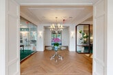 Located at 20013 Century Blvd in Germantown, Maryland, this beautifully appointed lobby serves as a welcoming first impression for the building. The entrance area is anchored by a striking herringbone-patterned hardwood floor that flows seamlessly throughout the space, lending a sense of refined elegance from the moment you step inside. A decorative round glass-top table with a polished silver base occupies the center of the foyer, accented by a vibrant floral arrangement that adds warmth and color. Overhead, a stylish multi-arm chandelier illuminates the space beneath a coffered ceiling adorned with detailed crown molding, reinforcing the classical architectural character. Flanking the entrance are glass-walled rooms — one housing a richly furnished lounge with deep emerald green carpet and curated artwork, and another serving as a dining or meeting area with complementary décor. Wall sconces provide soft ambient lighting throughout the hallway, while a built-in illuminated glass display case showcases decorative objects. A dedicated mailroom area along the hallway features a bank of individual secured mailboxes, offering convenient resident access. Natural light pours in through the exterior glass doors at the entrance, offering views of the surrounding outdoor greenery and creating an airy, inviting atmosphere throughout.