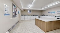 This shared laundry facility is a practical, well-organized space designed to accommodate high-volume use with efficiency and ease. The layout is open and spacious, providing comfortable room to navigate between machines and workstations without congestion.

A full row of top-load washing machines lines one wall, accompanied by a set of front-load dryers, offering users a variety of laundry options. A centrally positioned island unit with a wood-paneled base and a clean white countertop provides a convenient folding and sorting surface, complemented by an additional built-in counter along the perimeter wall — ideal for managing multiple loads simultaneously.

The flooring throughout is a classic speckled terrazzo, which is both durable and easy to maintain. Overhead fluorescent strip lighting ensures the space is consistently and evenly illuminated. Walls are finished in a neutral warm white, contributing to a clean, hygienic atmosphere. A small window provides a touch of natural ventilation, while baseboard heating keeps the space comfortable year-round.

Informational notices and instructional postings are mounted on the walls, keeping users informed on machine operation and building guidelines. A payment kiosk is also present, enabling convenient card-based access to the machines. The facility is accessible via a dedicated corridor and is equipped with a fire safety system, underscoring its commitment to user safety.