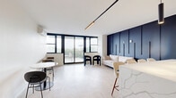 This thoughtfully designed 2-bedroom, 1-bathroom residence at 440 Elizabeth Ave in Newark, NJ offers a sleek, modern aesthetic throughout. The open-concept kitchen and living area serves as the heart of the home, anchored by a striking waterfall-edge quartz island with bar seating. The kitchen is outfitted with stainless steel appliances, warm wood lower cabinetry, white upper cabinets, and a marble-look tile backsplash, all illuminated by dramatic pendant track lighting. A bold navy accent wall with vertical paneling adds depth and character to the living space, which features light hardwood flooring and floor-to-ceiling black-framed windows and sliding glass doors leading to a private balcony with open outdoor views. The primary bedroom is generously proportioned, benefiting from large windows that fill the room with natural light, light hardwood floors, and a modern geometric ceiling fixture. The second bedroom offers a comfortable, quiet retreat with wall-to-wall carpet and a split-system air conditioning unit. The bathroom is elegantly finished with large-format tile, a soaking tub, textured olive-green subway tile surround, matte black fixtures, and a backlit mirror. Mini-split HVAC units are present throughout for individual climate control.