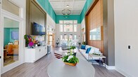 This single-floor clubhouse space offers a generously sized, coastal-inspired interior designed for both socializing and relaxation. The most striking architectural feature is the double-height ceiling in the main lounge area, accented by bold teal upper walls and illuminated by a pair of stacked circular pendant lights. Tall windows along the rear wall flood the space with natural light, creating an airy, welcoming ambiance throughout.

The open-plan layout connects several distinct functional zones. The main lounge features light hardwood-style flooring, a large sectional sofa with coordinating accent chairs, a vibrant area rug, and an organic-shaped coffee table. A wall-mounted large-format TV with a "Beachline" neon sign anchors a service counter equipped with a wine refrigerator and beverage station.

A separate lounge area incorporates a foosball table, casual seating clusters, and colorful artwork, continuing the playful coastal theme with teal ceilings and wood-paneled accents. The kitchen features white shaker cabinetry, a chevron tile backsplash, stainless steel appliances including a full refrigerator and dishwasher, open shelving, and a long communal dining table — ideal for group gatherings.