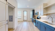 This single-floor unit offers a thoughtfully designed layout that maximizes its compact footprint. The open living and kitchen area is the centerpiece of the space, featuring a striking two-tone kitchen with bold navy-blue lower cabinetry contrasted by crisp white upper cabinets. A geometric tile backsplash, light quartz countertops, and a full suite of stainless steel appliances—including a refrigerator, range, microwave, and dishwasher—complete the functional and stylish kitchen. Light wood-style flooring runs throughout the main living areas, enhancing the bright, airy atmosphere created by recessed lighting and multiple windows that invite natural light. A sliding barn door adds a modern architectural detail, leading to additional spaces within the unit. The living area includes a built-in navy cabinet beneath a window, offering both storage and visual continuity with the kitchen's design palette. Two bathrooms serve the unit, one of which features a soaking tub and shower combination dressed in large-format marble-look tile with a decorative mosaic border accent, paired with overhead cabinetry for storage. A dedicated closet provides additional organization. The overall ambiance is clean, contemporary, and efficiently designed.