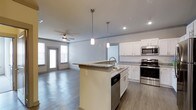 This single-story residence at Arbor Crossing offers an open-concept design that seamlessly connects the kitchen and living areas, creating a bright and functional living environment. The kitchen serves as the centerpiece of the space, anchored by a central island topped with granite countertops and equipped with an undermount sink and pendant lighting above. White cabinetry lines the perimeter walls, complemented by matching granite countertops and a full suite of stainless steel appliances, including a gas range, microwave, and dishwasher. Recessed lighting throughout the kitchen ensures ample illumination.

The open layout flows naturally into the living area, where wood-look flooring extends seamlessly across both spaces, adding warmth and visual continuity. A ceiling fan provides practical comfort in the living room, which benefits from natural light through multiple windows. Neutral gray wall tones throughout create a clean, contemporary aesthetic that pairs well with the light-toned flooring. A set of doors at the far end of the living area provides access to the exterior, enhancing the sense of openness and connectivity to the outdoors.