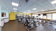 This fitness center is designed to support a well-rounded workout experience within a single open floor layout. The space is outfitted with a solid selection of cardio equipment, including elliptical trainers, upright and recycle bikes, treadmills, and rowing machines, all arranged to allow comfortable circulation between stations. The flooring consists of dark, durable rubber matting suited for high-traffic fitness use.

A large mirrored wall runs along one side of the room, visually expanding the space and allowing users to monitor their form during workouts. The walls feature bold yellow accents that add energy to the environment. A wall-mounted flat-screen television is positioned for easy viewing from the cardio equipment.

Natural light enters through a row of windows along one wall, complementing the bright, recessed panel lighting overhead set within a drop ceiling. A wall clock is also visible, providing practical utility for timed workouts. The overall ambiance is functional and motivating, offering a clean, organized environment with the essential equipment needed for a consistent fitness routine.