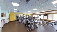 This fitness center is designed to support a well-rounded workout experience within a single open floor layout. The space is outfitted with a solid selection of cardio equipment, including elliptical trainers, upright and recycle bikes, treadmills, and rowing machines, all arranged to allow comfortable circulation between stations. The flooring consists of dark, durable rubber matting suited for high-traffic fitness use.

A large mirrored wall runs along one side of the room, visually expanding the space and allowing users to monitor their form during workouts. The walls feature bold yellow accents that add energy to the environment. A wall-mounted flat-screen television is positioned for easy viewing from the cardio equipment.

Natural light enters through a row of windows along one wall, complementing the bright, recessed panel lighting overhead set within a drop ceiling. A wall clock is also visible, providing practical utility for timed workouts. The overall ambiance is functional and motivating, offering a clean, organized environment with the essential equipment needed for a consistent fitness routine.