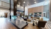 Located at 2902 N Arthur Ashe Blvd in Richmond, Virginia, this expansive single-floor amenity lounge offers a generously sized, open-plan layout designed with a sophisticated blend of modern and industrial aesthetics. Soaring double-height ceilings amplify the sense of space throughout, while warm wood-look tile flooring and exposed brick accent walls provide a grounded, inviting atmosphere.

A striking centerpiece double-sided glass fireplace, framed in dark glazed tile and topped with a mounted flat-screen display, anchors the main lounge area. Multiple distinct seating zones feature plush, contemporary sectionals and accent chairs in rich earth tones, arranged to encourage both social gatherings and relaxed lounging. A built-in bookshelf nook offers a cozy library-style retreat.

Toward the rear, a fully appointed kitchen and dining area showcases dark cabinetry, open shelving stocked with dishware, a large communal dining table, and a dramatic living green wall that cascades lush foliage across the ceiling. A bar counter with seating provides an additional gathering point. A wide corridor lined with curated wall art and leather lounge chairs connects the zones, while views into an adjacent fitness area and an upper-level mezzanine add depth and dimension to this thoughtfully designed communal space.