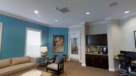 This single-floor space offers a thoughtfully designed interior that balances comfort and functionality. The main area serves as an inviting lounge, featuring a rich teal accent wall adorned with a colorful floral artwork that adds a vibrant focal point to the room. Neutral taupe tones on the surrounding walls complement the bold color, creating a well-balanced and welcoming ambiance. Seating includes a stylishly crafted wooden chair with a cushioned seat and a coordinating side table with a classic table lamp, providing a comfortable and intimate setting.

The space is finished with elegant crown molding along the ceiling, which is equipped with recessed lighting that ensures even, bright illumination throughout. Herringbone-patterned hardwood flooring adds a classic touch of texture underfoot. Natural light filters in through a window fitted with horizontal blinds.

Adjacent to the lounge area is a well-appointed refreshment station, featuring dark espresso-finished cabinetry with ample storage, granite countertops, and a coffee and beverage setup. A glass-paneled interior door connects to an additional area beyond, maintaining an open flow while providing separation between spaces. Overall, the design is cohesive, professional, and inviting.
