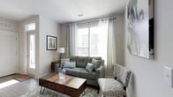 This single-floor residence offers a thoughtfully designed living space that balances comfort and contemporary style. The living room serves as the heart of the home, centered around a tufted gray sofa complemented by patterned accent pillows and a coordinating upholstered armchair. A substantial wooden coffee table with hairpin legs and built-in drawers anchors the seating arrangement, providing both a focal point and practical storage. Natural light pours in through a large window dressed with sheer white curtains and gray draping panels, creating a bright and airy atmosphere. The neutral color palette — soft grays, warm whites, and earthy browns — lends the space a calm, cohesive feel. A wooden side table with a sculptural lamp adds warmth alongside framed wall art that enhances the curated aesthetic. The flooring features a patterned area rug that defines the seating zone and adds texture underfoot. Clean white walls and a smooth ceiling with a centrally placed ventilation fixture reflect the space's clean-lined, modern sensibility. Overall, the residence presents a welcoming and functional environment with attention to detail in both furnishings and layout.
