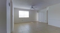 This single-story space features an open living area finished with light-toned ceramic tile flooring that extends throughout, lending a clean and cohesive look to the interior. The room is generously sized, offering a comfortable sense of openness suited for a variety of furniture arrangements. Natural light enters through a large multi-pane window along one wall, while a set of sliding glass doors fitted with vertical blinds provides additional brightness and direct access to the outdoors. A ceiling fan is centrally mounted overhead, contributing both to air circulation and a functional aesthetic. The walls are painted in a neutral white tone, complementing the light flooring and creating a bright, airy atmosphere. The ceiling features a textured popcorn finish, typical of the construction style. Multiple electrical outlets are positioned along the baseboards, offering practical convenience throughout the space. Overall, the living area presents a straightforward, well-lit interior with functional features well-suited for everyday living.