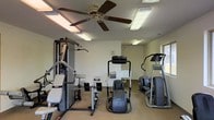 This single-floor fitness center offers a practical and functional workout space designed to accommodate a range of exercise needs. The room is outfitted with a selection of strength training equipment, including a multi-station cable and weight machine, as well as dedicated seating for isolated exercises. For cardiovascular training, the space features an elliptical trainer and a stair climber, providing users with effective aerobic workout options. A ceiling fan anchors the center of the room, working alongside multiple overhead fluorescent light fixtures to maintain a bright, well-ventilated atmosphere throughout. The walls are finished in a neutral tone, contributing to a clean and focused environment. A water fountain is conveniently positioned along one wall, ensuring easy hydration access during workouts. Natural light is introduced through a window on the far wall, adding to the overall brightness of the space. The carpeted flooring provides a comfortable underfoot surface, while the open layout allows for easy navigation between equipment. Overall, this fitness center delivers a straightforward and well-maintained space suited for both strength and cardio training routines.