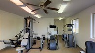 This single-floor fitness center offers a practical and functional workout space designed to accommodate a range of exercise needs. The room is outfitted with a selection of strength training equipment, including a multi-station cable and weight machine, as well as dedicated seating for isolated exercises. For cardiovascular training, the space features an elliptical trainer and a stair climber, providing users with effective aerobic workout options. A ceiling fan anchors the center of the room, working alongside multiple overhead fluorescent light fixtures to maintain a bright, well-ventilated atmosphere throughout. The walls are finished in a neutral tone, contributing to a clean and focused environment. A water fountain is conveniently positioned along one wall, ensuring easy hydration access during workouts. Natural light is introduced through a window on the far wall, adding to the overall brightness of the space. The carpeted flooring provides a comfortable underfoot surface, while the open layout allows for easy navigation between equipment. Overall, this fitness center delivers a straightforward and well-maintained space suited for both strength and cardio training routines.