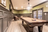 Located on Campbell Street in Rahway, New Jersey, this single-level billiards and game room is designed with a refined, club-inspired aesthetic. The centerpiece of the space is a full-size pool table with a rich mahogany finish and cream felt surface, positioned beneath a striking oversized pendant light with a metallic disc shade that provides focused illumination over the playing surface.

The room's design is defined by its bold two-tone wall treatment: dark charcoal wainscoting with detailed raised-panel molding lines the lower half of every wall, while a muted sage green paint carries to the ceiling, creating a sophisticated, moody atmosphere. Crown molding runs continuously along the upper perimeter, adding a polished architectural finish.

A dedicated lounge area along one wall features a grouping of brown leather barrel chairs arranged around a central table on a textured area rug, offering a comfortable space for spectators and socializing. Card-themed framed artwork decorates the walls, reinforcing the playful yet upscale character of the room. A wall-mounted flat-screen television, a cue rack, and sconce lighting complete the amenities. Light wood-toned flooring runs throughout, contrasting warmly against the dark wainscoting. French-style glass doors provide access to adjacent areas, allowing natural light to filter into the space.