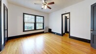 This single-floor unit carries a consistent design language throughout, defined by warm honey-toned hardwood floors and striking black door frames, trim, and baseboards that create strong visual contrast against white walls. The living area is a comfortable, open space bathed in natural light from a pair of large windows dressed with blinds. A ceiling fan with integrated lighting overhead and a classic cast-iron radiator beneath the windows round out the room's functional character.

The bedroom offers two separate openings—one leading to a dedicated closet fitted with a hanging rod, and another connecting to an adjacent space, providing flexible storage and layout options. The kitchen is equipped with warm oak cabinetry spanning both upper and lower sections, a stainless steel sink with a light countertop, and a full-size white refrigerator. Tile flooring in this area transitions from the hardwood found elsewhere.

The bathroom maintains a clean, classic aesthetic with white penny-tile flooring, white wall tiles, a pedestal sink, a soaking tub with a shower curtain rod, and a window that allows natural light into the space. Black door hardware and framing carry the unit's consistent design theme throughout.