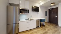 Located at 78 Dorrance St in the heart of Providence, Rhode Island, this single-floor unit blends modern design with character-rich architectural details. Wood-look flooring runs consistently throughout the space, lending warmth and continuity to the interior.

The bedroom offers a clean, contemporary aesthetic, anchored by a textured chevron-patterned feature wall that adds visual depth. A wall-mounted flat-screen television, built-in storage bed frame, and a bright window overlooking the urban streetscape complete the room's functional yet stylish layout.

The kitchenette is efficiently designed with sleek white cabinetry, a crisp white countertop, induction cooktop, stainless steel refrigerator, over-range microwave, and a single-basin sink. A wall-mounted television and integrated workspace with seating make this area multifunctional.

The bathroom is finished with large-format grey stone-look tiles covering both the walls and floor, creating a refined, spa-like atmosphere. A vessel sink atop a white vanity, illuminated mirror cabinet, and modern chrome fixtures add a polished touch.

A notable feature visible in the layout is an exposed brick interior wall, offering a nod to the building's historic character while contrasting beautifully with the otherwise contemporary finishes throughout.