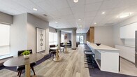 Located at 851 Richmond Rd in Ottawa's vibrant Westboro neighbourhood, this thoughtfully designed amenity lounge offers a generous, open-concept layout across a single level. The space is organized into several distinct functional zones that flow seamlessly into one another, creating an inviting and versatile environment.

Upon entry, a glass-fronted vestibule with automatic doors leads into the main area, where patterned carpet tiles in neutral tones cover much of the floor, complemented by sections of hard flooring along the perimeter. Structural columns punctuate the space while adding to its architectural character.

The lounge area features multiple seating arrangements, including leather armchairs and a plush sofa grouped around a low coffee table, as well as bistro-style tables paired with upholstered chairs — ideal for casual conversations or quiet relaxation. Large windows fitted with roller shades allow diffused natural light to fill the interior throughout the day.

A standout feature is the long kitchenette bar, finished with a crisp white countertop and equipped with a sink, bar stools, and warm wood cabinetry with open shelving above. A stainless steel refrigerator and wall-mounted television complete this practical yet stylish area.

Recessed pot lighting in a suspended tile ceiling ensures consistent, even illumination throughout. Curated wall art and live greenery add warmth and personality to the overall ambiance.