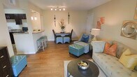 This single-floor residence offers a thoughtfully designed open-concept layout that seamlessly connects the living, dining, and kitchen areas into one inviting, cohesive space. Rich hardwood flooring flows throughout, adding warmth and continuity to the interior.

The living area is appointed with elegant tufted seating in soft neutral tones, complemented by a teal ottoman that serves as a stylish focal point. A round dark-metal coffee table anchors the seating arrangement, while a large area rug defines the space beneath.

The dining area features a classic dark wood table with cushioned seating and teal upholstered ottomans, illuminated by a sleek track lighting fixture overhead. Decorative wall sconces flank a tasteful floral arrangement, adding a refined, curated touch to the space.

Adjacent to the dining area, the kitchen pass-through features a raised breakfast bar with bar stools, allowing for casual dining and easy interaction between the kitchen and living spaces. Pendant lighting above the bar adds a warm, inviting glow.

The overall ambiance is bright and airy, with neutral-toned walls providing a clean backdrop that allows the carefully chosen furnishings and accent colors — including coral and teal — to stand out. The layout is efficient yet spacious, offering a comfortable and stylish living environment.