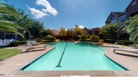 This outdoor amenity space centers around a generously sized swimming pool with clear turquoise water, accented by a defined lap lane. The pool deck is finished in a neutral concrete surface and is surrounded by neatly trimmed landscaping, including mature palm trees, ornamental shrubs, and flowering plants that contribute to a resort-like atmosphere.

Multiple seating areas are distributed throughout the space to accommodate varied preferences. A prominent gazebo with a bold orange canopy roof provides shaded relief from the sun, furnished with wicker lounge chairs and a sofa with navy blue cushions, creating a comfortable retreat adjacent to the pool. Additional chaise lounges are positioned along the pool's edge for sunbathing.

A secondary shaded seating area features a canopy structure with plush blue cushioned furniture, offering views of the surrounding greenery and community buildings. The landscaping throughout includes tall privacy trees, manicured hedges, and potted tropical plants that enhance the visual appeal of the area.

The space is enclosed by decorative iron fencing, providing both security and an open sightline to the surrounding grounds. Brick and multi-story residential buildings frame the background, complementing the overall aesthetic of the community.