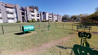 This community amenity space offers two distinct areas tailored to both residents and their four-legged companions. The outdoor dog park is a generously sized, fully enclosed green space secured by chain-link fencing, providing a safe environment for pets to roam freely. Agility features, including a tunnel and interactive equipment, are thoughtfully placed throughout the grassy grounds, set against a backdrop of mature trees and a clear open sky.

The fitness center is a well-appointed, single-level space distinguished by a bold color scheme featuring orange and blue accent stripes along the upper walls, lending the room a vibrant, energetic atmosphere. Patterned carpet flooring covers the workout area, which is equipped with multiple treadmills, upright and recumbent stationary bikes, an elliptical trainer, a multi-station cable machine, and a full dumbbell rack. A large wall-mounted mirror amplifies the sense of space and aids in form monitoring during workouts. Recessed lighting and a ceiling fan ensure a comfortable environment, while several windows allow abundant natural light and views of the outdoor pool area and surrounding grounds. A glass-paneled entry door and clearly posted safety equipment complete the functional, well-maintained space.