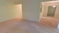 This single-floor space features an open layout with wall-to-wall carpet in a warm neutral tone, creating a cohesive and comfortable feel throughout. The main living area is generously sized, with smooth white walls and a central architectural dividing wall that subtly separates the space into distinct zones while maintaining an open, connected atmosphere. A recessed opening on the left side of the dividing wall leads to an additional carpeted room, providing flexible use as a bedroom, office, or secondary living area. To the right, a hallway connects to a kitchen area, where a countertop pass-through is visible, offering a convenient link between the kitchen and living spaces. Beyond the hallway, a door leads to what appears to be a bathroom or additional room, contributing to the overall functionality of the layout. The uniform carpeting and consistent neutral color palette throughout lend the space a clean, understated aesthetic. Natural light filters in from unseen windows, brightening the interior. The straightforward, practical design makes this space highly adaptable for a variety of furnishing arrangements and lifestyle needs.